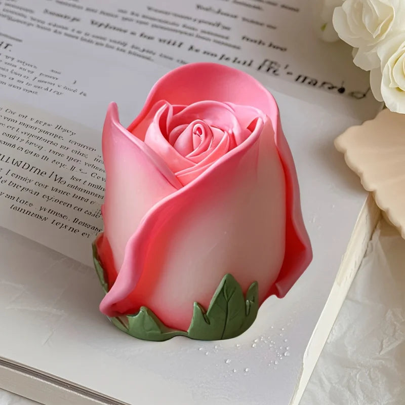 A pink rose silicone candle mold with green leaves resting on a white surface next to some paper.