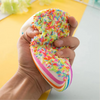 A hand holds a rainbow layer cake squishy toy covered in colorful sprinkles against a yellow background.
