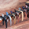 A row of professional metal guitar capos with pick holders in various colors and finishes resting on a wooden surface.