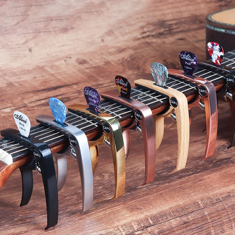 A row of professional metal guitar capos with pick holders in various colors and finishes resting on a wooden surface.
