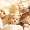 A close-up of a lifelike 14-inch reborn baby doll with blonde hair and a white outfit lying on a soft, light-colored fabric.