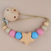 A wooden star shaped pacifier clip with silicone beads and a leather cord on a light pink background.