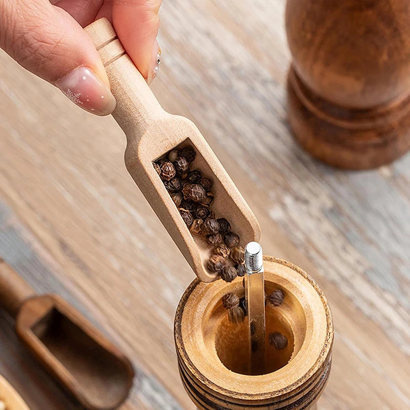A hand is using a wooden scoop spoon to extract peppercorns from a wooden grinder.