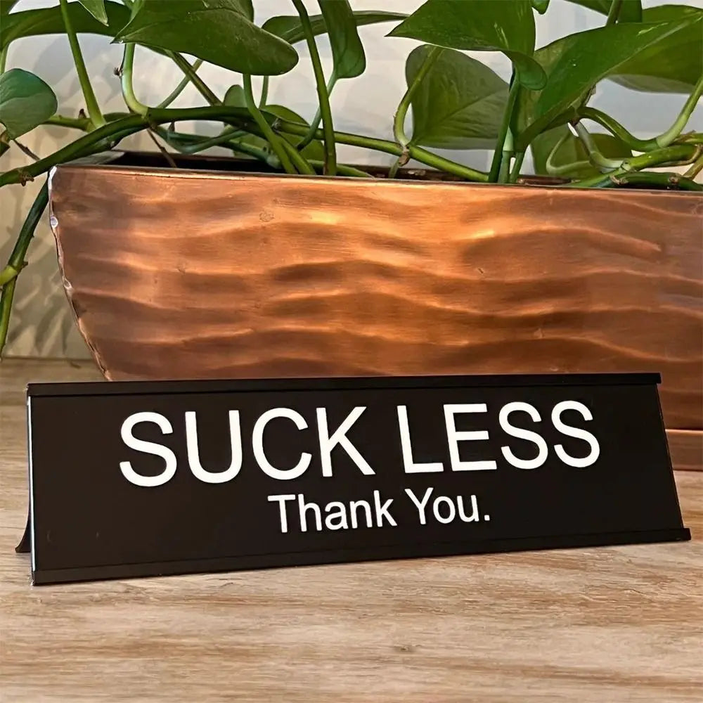 A black desk sign with white text that reads Suck Less Thank You placed in front of a potted green plant.