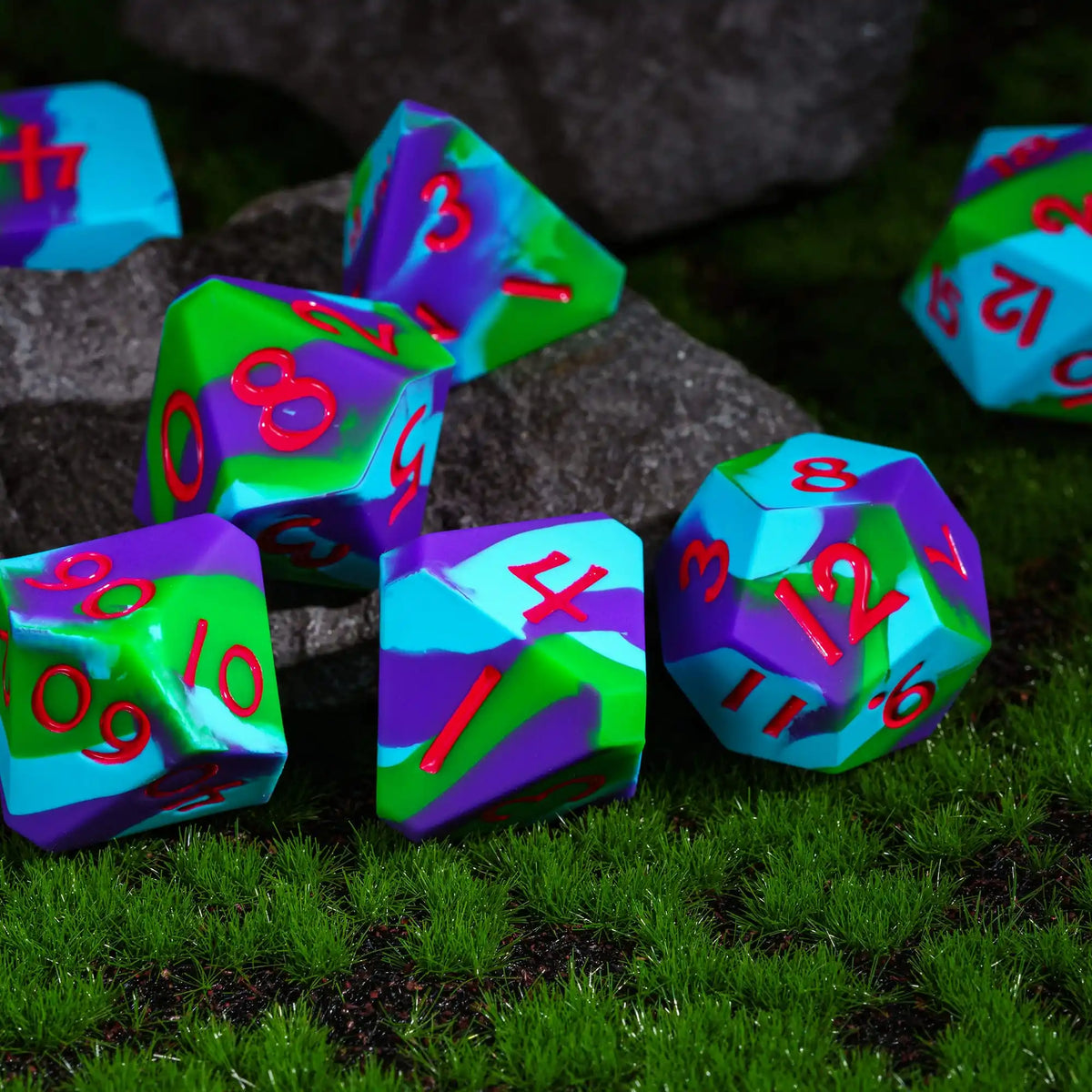 A collection of colorful silicone polyhedral dice resting on green grass and dark rocks.