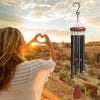 A woman holds her hands in a heart shape against a sunset backdrop while a wind chime hangs nearby.