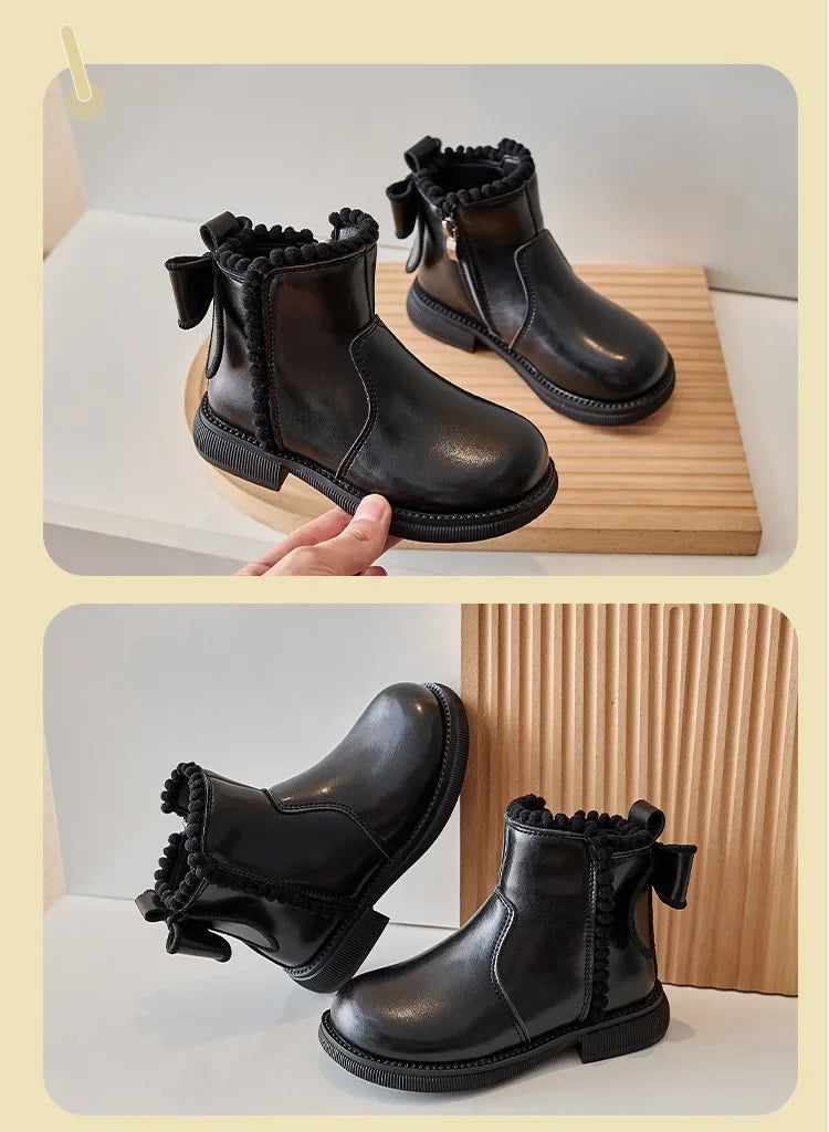 A pair of black girls' ankle boots with a bow-knot detail and side zipper displayed on a wooden surface.