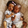 A newborn twin baby doll with dark skin is lying on a fluffy beige surface.