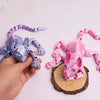 A hand holds a purple and silver articulated 3D printed sphynx cat figurine with a pink, snake-like tail.