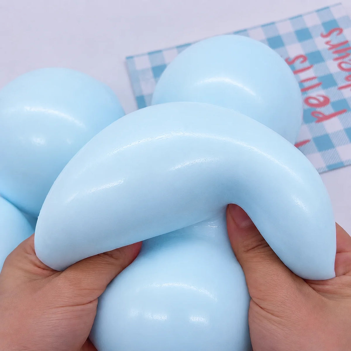 A hand is squeezing a light blue slow rising squishy ball stress relief fidget toy against a checkered background.