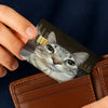 A hand holds a card featuring a cat face meme design with a gold sticker on a brown leather wallet.