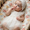 A newborn baby girl wearing a white lace tulle dress and hat, lying in a wicker bassinet.