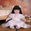 A lifelike reborn baby doll with soft cloth body, dressed in a floral white dress, sitting on a wooden surface.