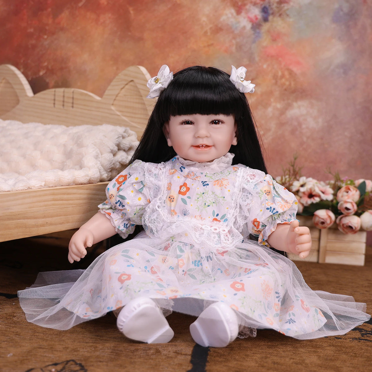 A lifelike reborn baby doll with soft cloth body, dressed in a floral white dress, sitting on a wooden surface.