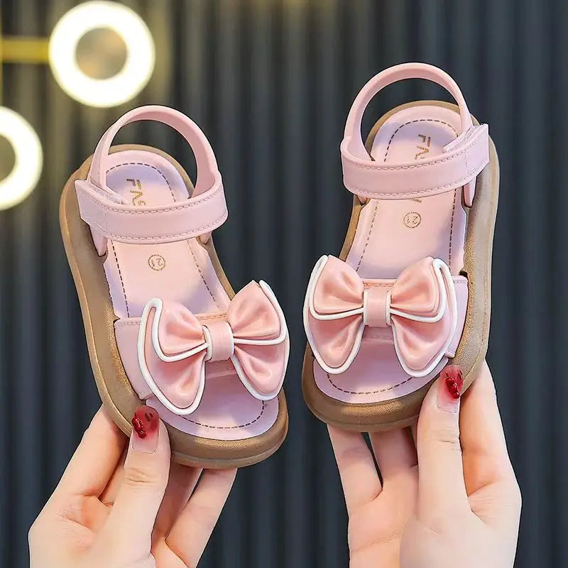 Pair of pink girls summer sandals with bow details being held by hands.