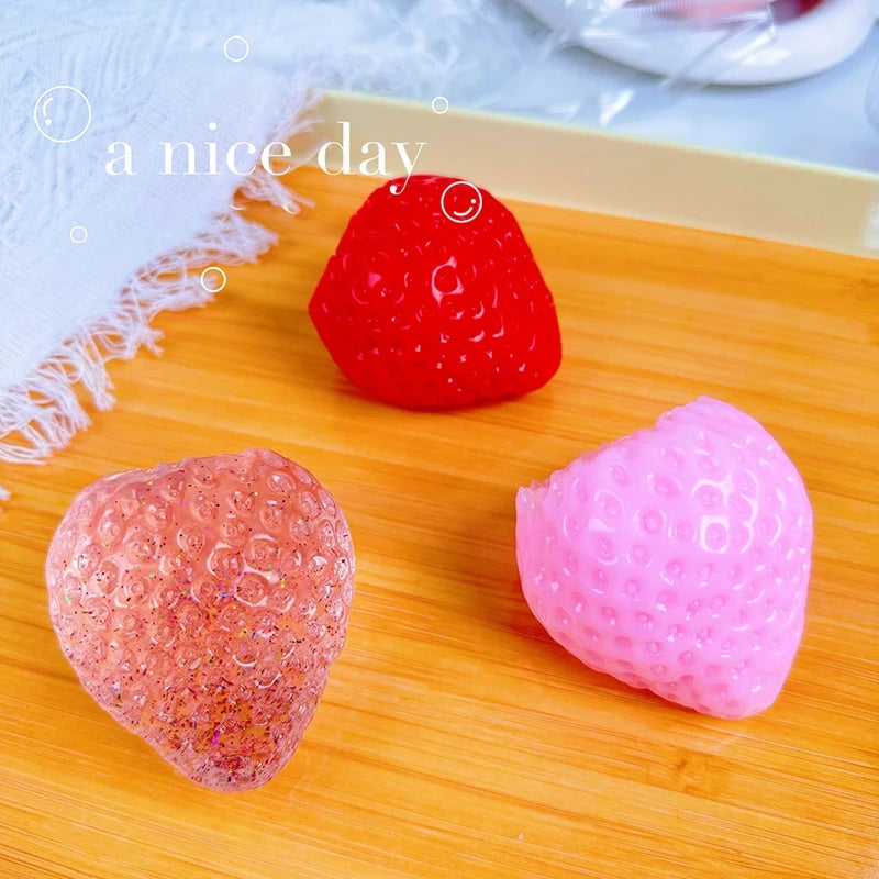 Two squishy strawberry fidget toys, one red and one pink, are displayed on a wooden surface.