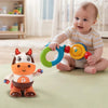 A baby is interacting with an orange and yellow musical cow plush toy that has a green ring attached.