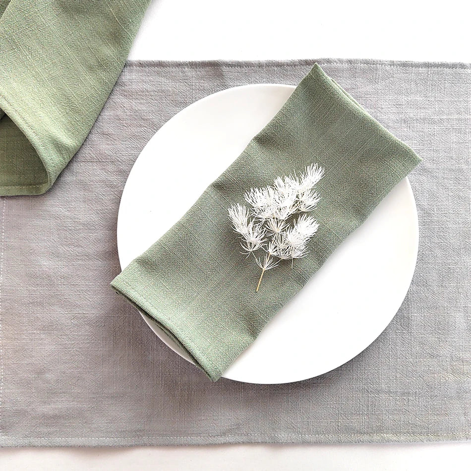 A stack of olive green cotton-linen dinner napkins placed on a gray linen placemat with a white plate.