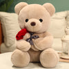 A light beige teddy bear plush toy holding a red rose, sitting on a white surface.