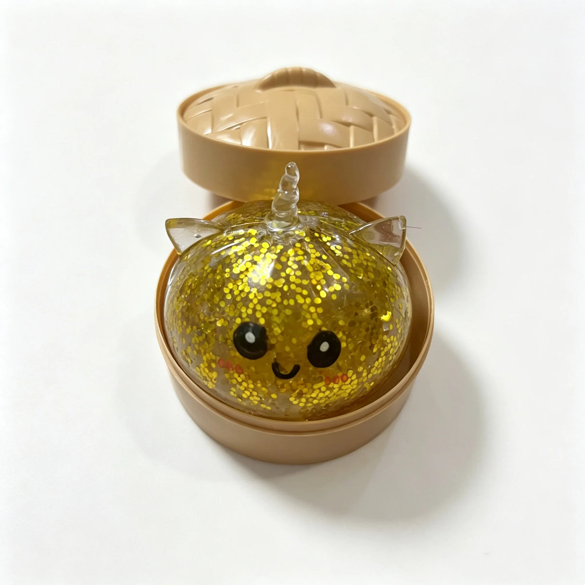 A golden glitter unicorn dumpling squishy stress ball is shown in its bamboo steamer box.