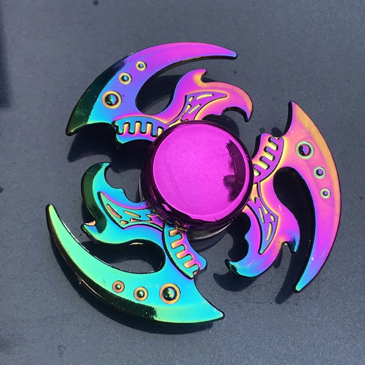A vibrant iridescent rainbow metal fidget spinner featuring a central sphere and surrounding wing-like blades.