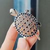 A close-up of a gold-toned rhinestone turtle brooch pin being held by a hand.