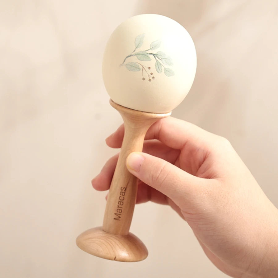 A hand holds a wooden maraca with a light-colored ball decorated with green leaves.