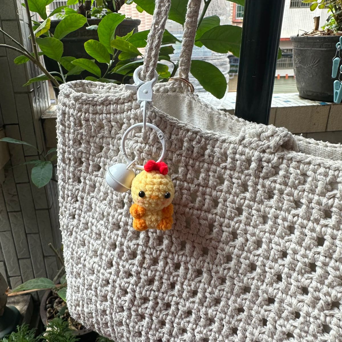 A handmade crochet animal keychain with a yellow knitted pendant and red flower detail attached to a cream-colored woven bag.