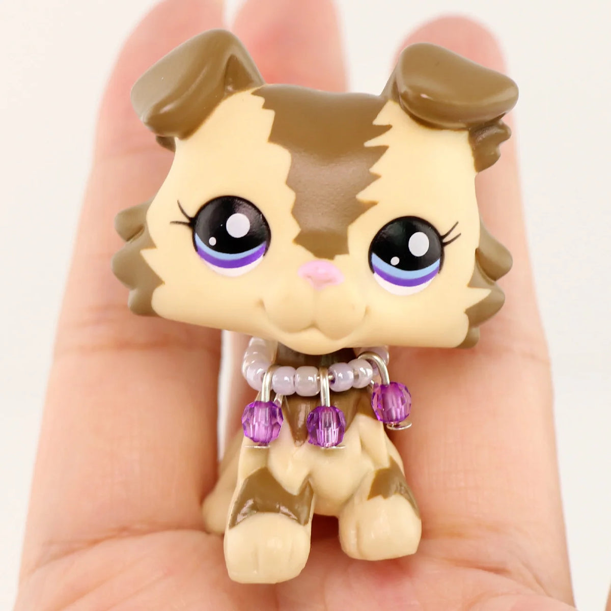 A miniature pet animal figure with brown and tan markings and purple bead accessories is held in a hand.