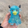 A hand holds a bright blue glitter Highland cow squishy stress relief toy with black horns and a yellow snout.