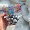 A clear plastic pouch containing a black and white cat paw squishy stress relief toy and a colorful packaging card.