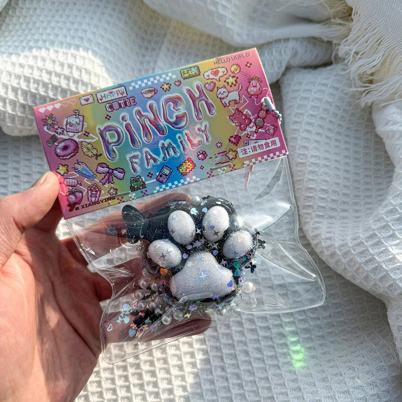 A clear plastic pouch containing a black and white cat paw squishy stress relief toy and a colorful packaging card.
