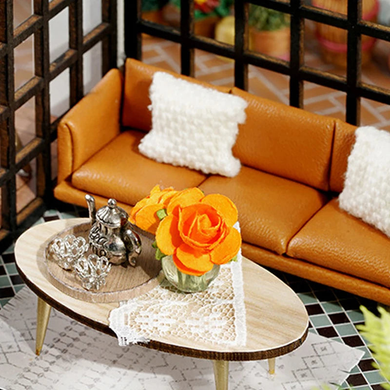 A miniature dollhouse puzzle kit displayed on a wooden table with an orange rose and white fluffy cushion.