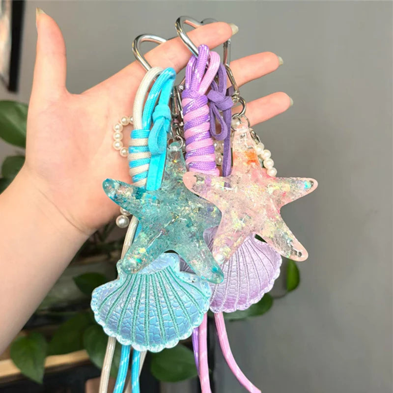 A hand holds a set of beaded keychains featuring starfish and seashell charms in shades of blue, purple, and pink.