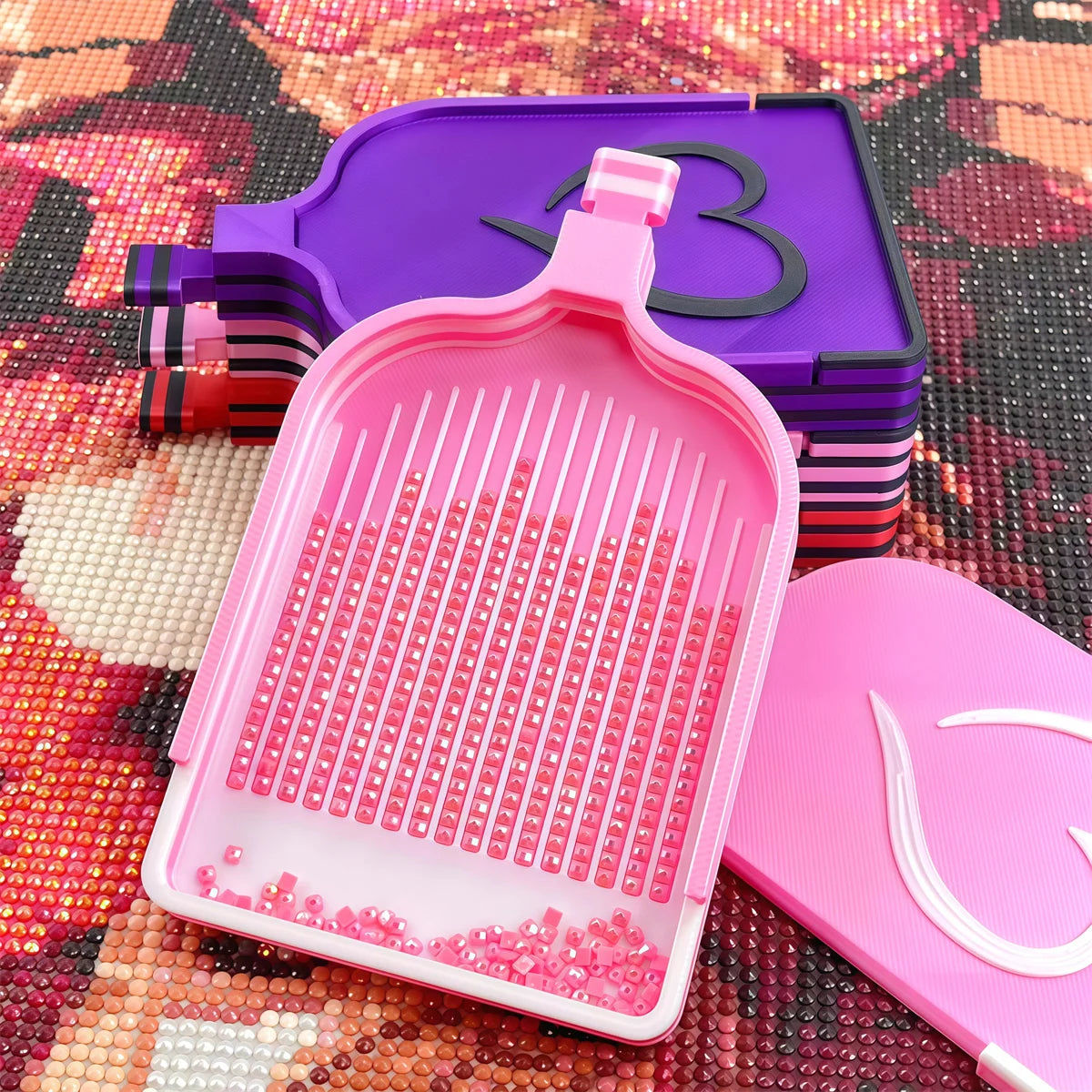 A set of diamond painting storage trays and organizer boxes in pink and purple are displayed on a textured surface.