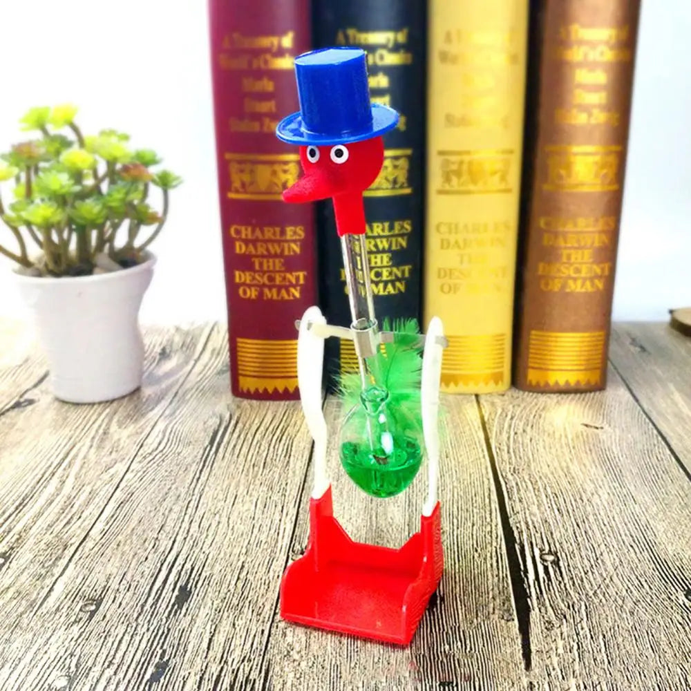 A red and blue bird toy with a long neck stands on a wooden surface next to books.
