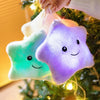 A hand is holding a glowing star-shaped plush pillow with an LED light, hanging on a Christmas tree.
