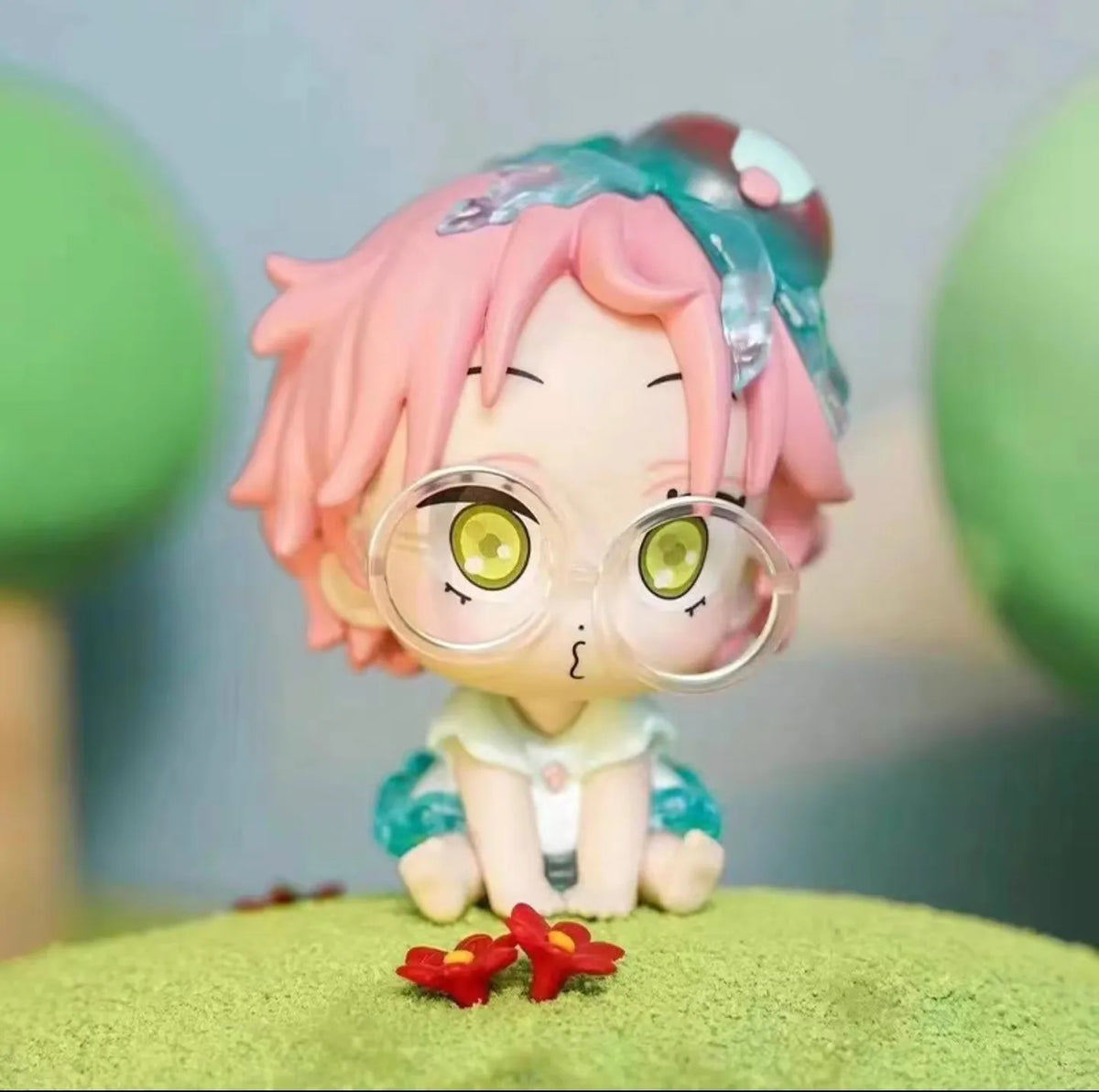 A mini figure collectible of a character with pink hair and green eyes wearing round glasses and blue clothing.