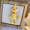 Handmade crochet frog hanging ornament with yellow body and pink accents displayed in a wooden frame