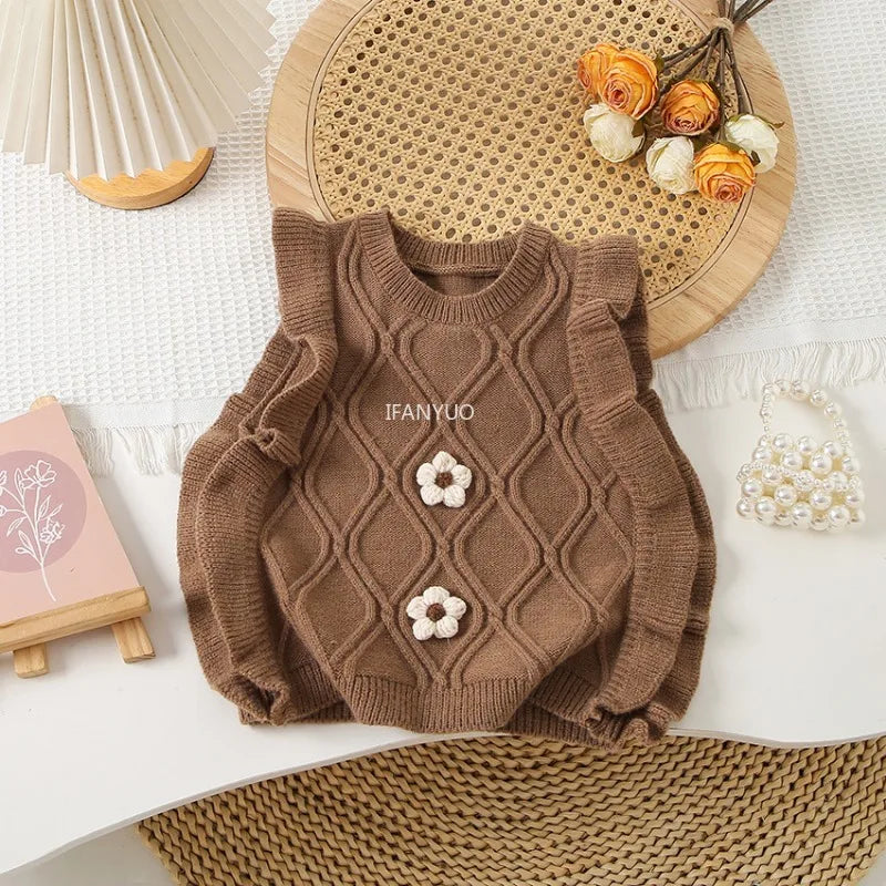 A brown knit sweater vest with a flower shape detail is displayed on a white surface next to a woven mat and dried flowers.