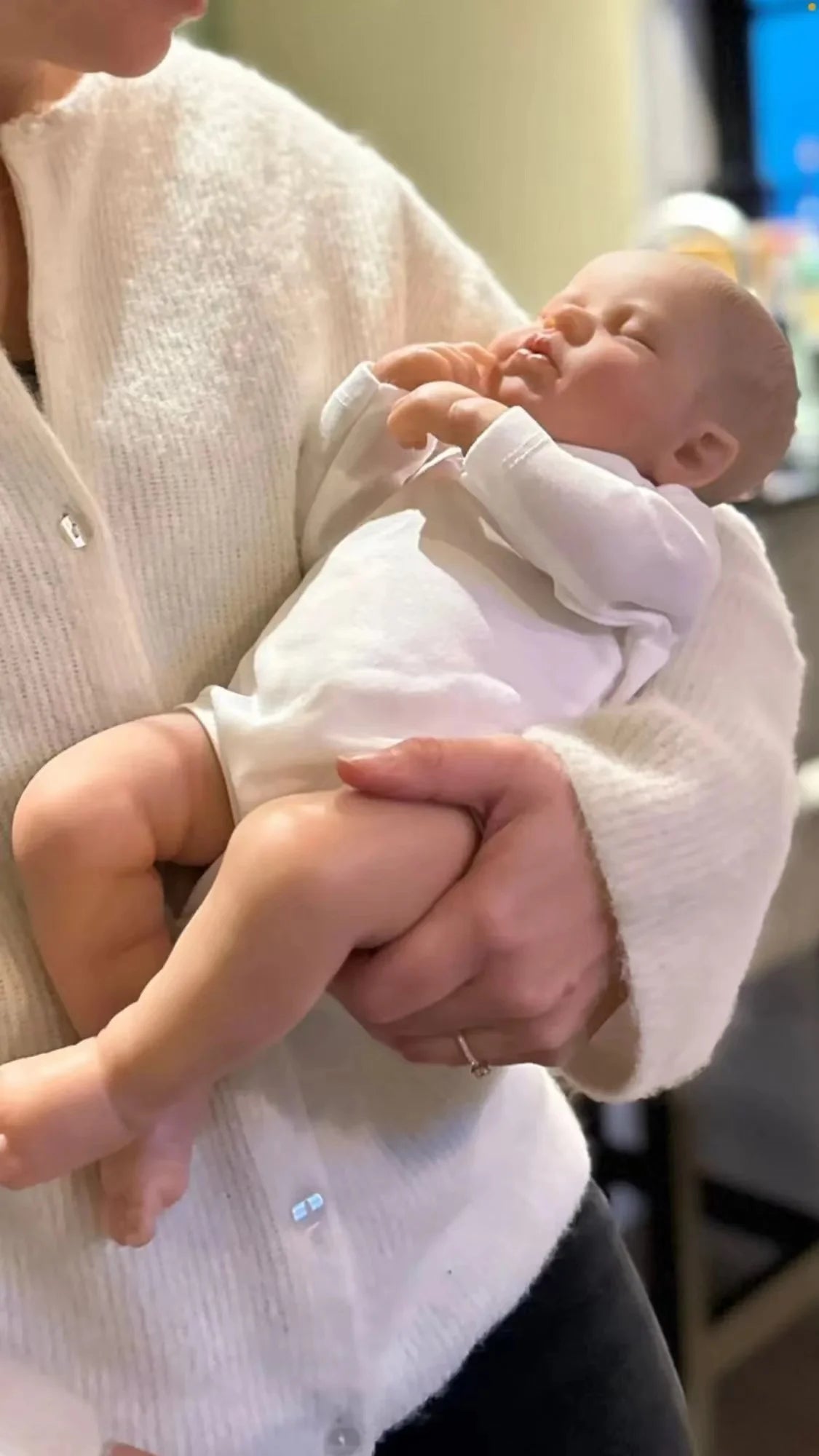 A person is holding a realistic reborn baby doll with a cloth body in a white knitted sweater.