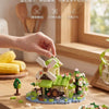 A hand is assembling a miniature forest cabin set featuring a greenhouse and windmill blocks on a wooden table.