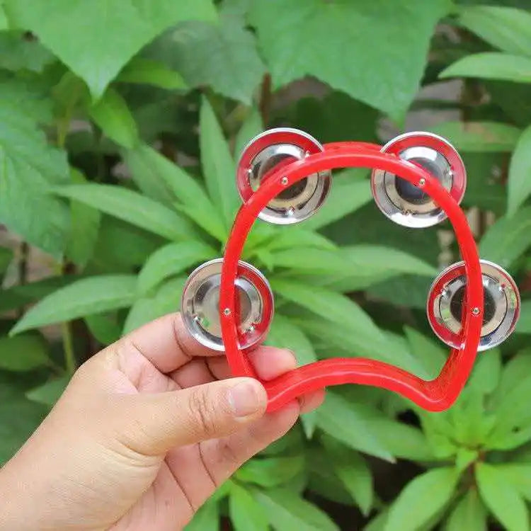 Una mano sostiene un tambor de percusión rojo portátil con cascabeles de plata contra un fondo de hojas verdes.