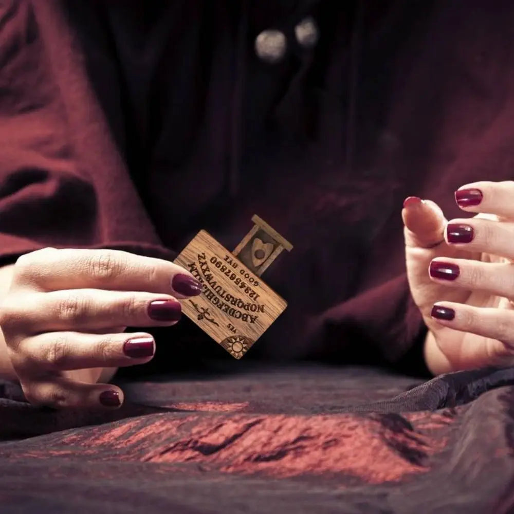 A hand holds a wooden miniature Ouija board resting on dark fabric.