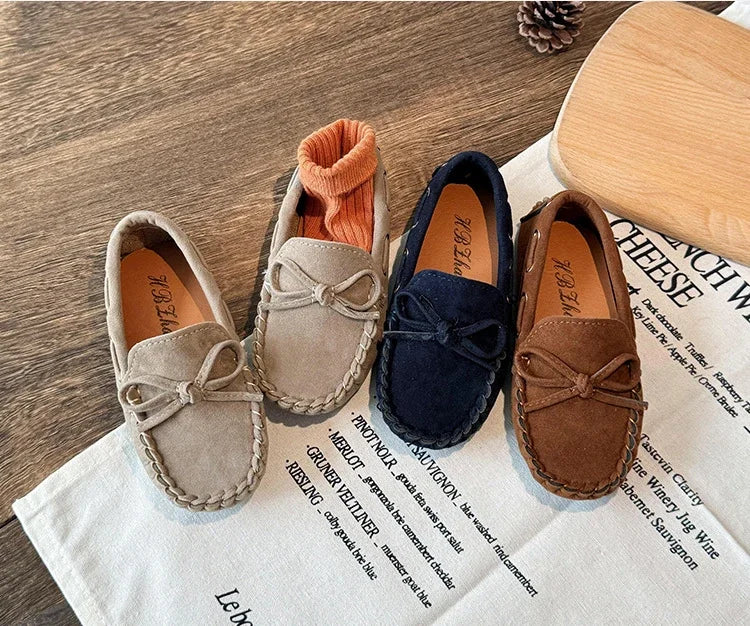 A pair of girls' brown slip-on moccasins with orange lining and bow details resting on a white cloth.