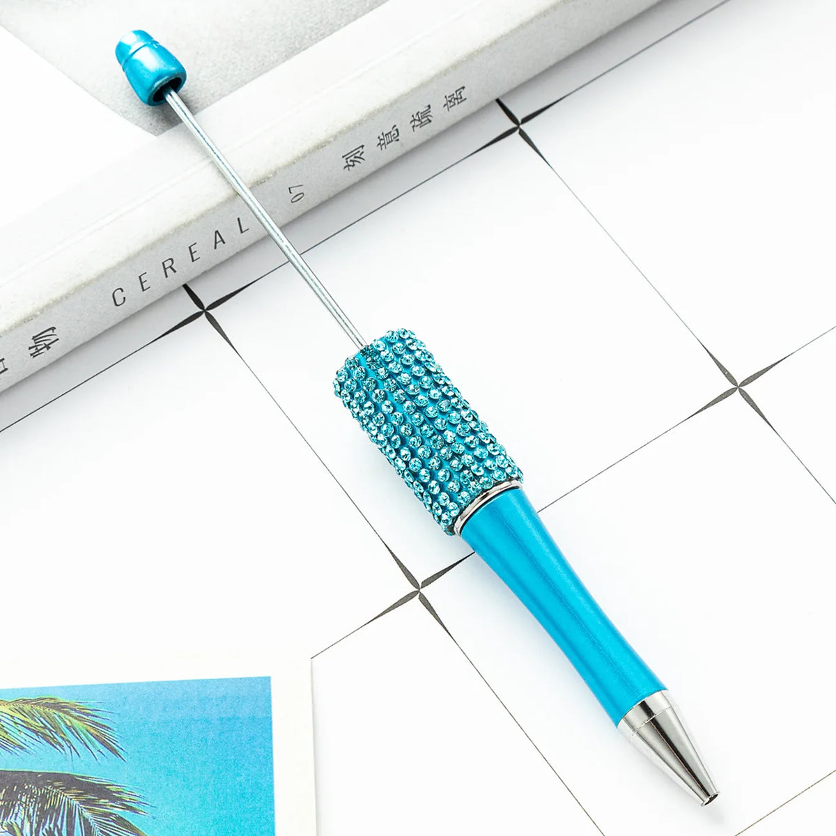 A blue beaded ballpoint pen craft kit with a silver tip rests on a white surface next to a book.