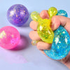 A hand holds a bright yellow squeeze toy with a green tip and a blue glittery duck-shaped fidget toy, next to a purple glitte