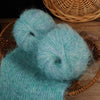 Two fluffy teal alpaca blend yarn balls resting on a woven basket and wooden surface.