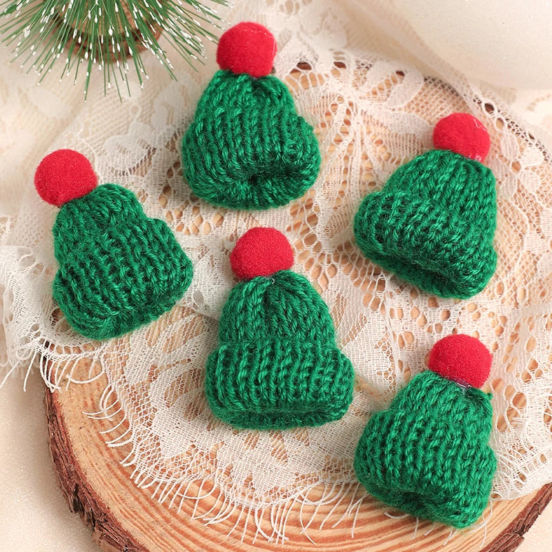 A set of mini knitted green Christmas doll hats with red pom-poms resting on a lace fabric and wooden surface.