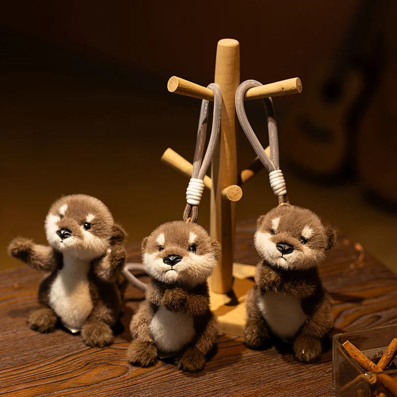 Three small brown otter plush toys hanging from a wooden cross keychain charm